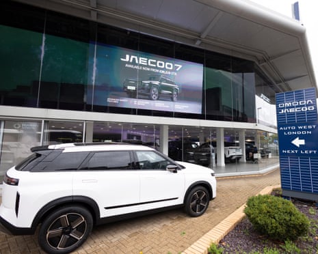 A white SUV is parked outside the Omoda/Jaecoo dealership, screen advertising "Jaecoo 7"
