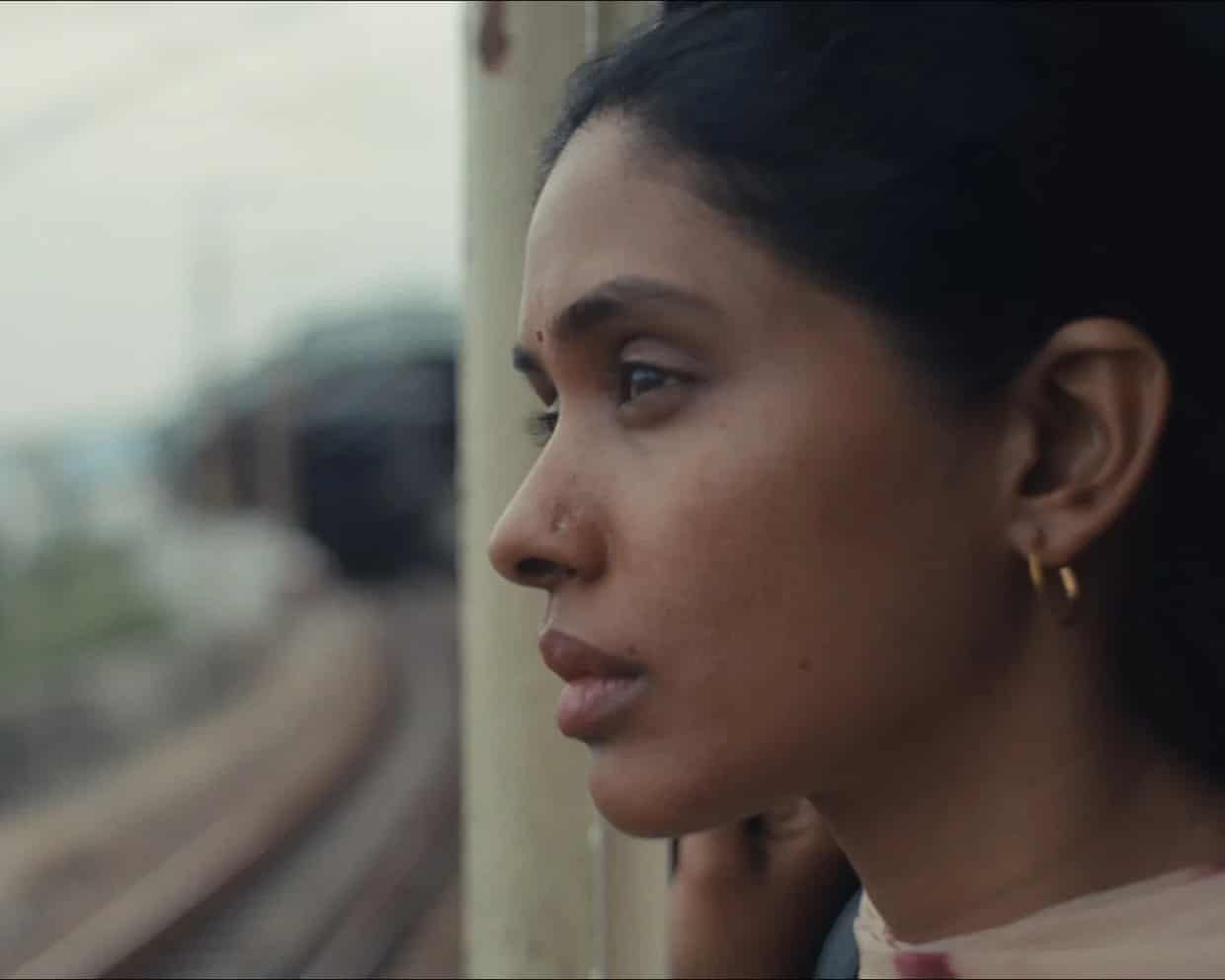 Selvi gazes out of a train window in profile, hand resting near her neck