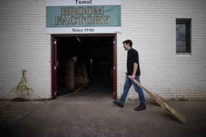 Man holding a broom in front of a brick wall and door, with the sign 'Tumut Broom Factory'