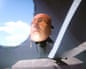 A synthetic human face is placed onto a machine, the whole head raised above a platform as the sky can be seen outside a window.
