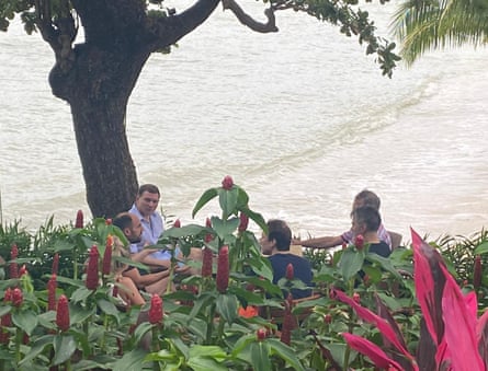 Five men and one woman sitting around a shore-side table near a tree, with luscious exotic plants in the foreground