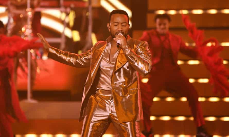 A man in a shiny golden suit performs on stage with a red backing dancer
