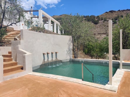 A small pool surrounded by terracotta coloured paving and steps and white walls with trees and a hill in the background