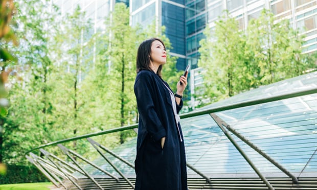 A confident young Asian woman looking at view, holding a smart phone, standing against corporate buildings at the park.