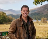 Matt Baker smiles in a brown jacket, standing outdoors with mountains and fields behind him