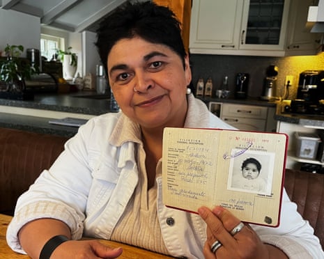 A middle-aged woman sits in a tidy kitchen and show off her passport from when she was an infant.