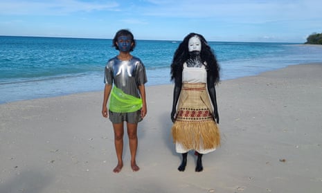 Two young women pose on a beach – one in a facepaint and a shiny metallic dress and the other in a mask and woven grass mat skirt