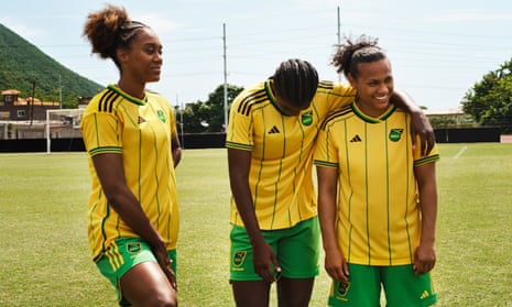 Three women standing on a football pitch wearing the Jamaica World Cup 2023 kit of yellow top with thin vertical green stripes, and green shorts