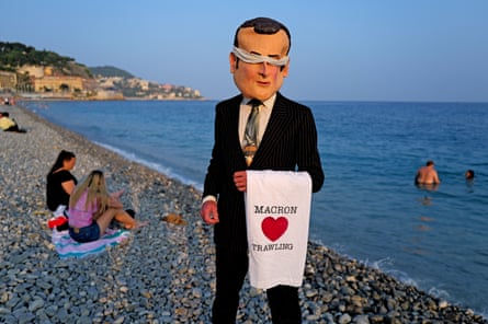 A person wearing a suit and a large papier-mâché head with its eyes covered stands of a beach holding a towel which reads ‘Macron hearts trawling’.
