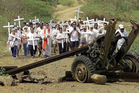 Crowd of people wearing white tops, carrying white crosses, with abandoned military vehicle in foreground