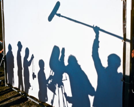 Shadows of a film crew at work are projected on a white screen, including people standing with cameras and holding a high boom mic