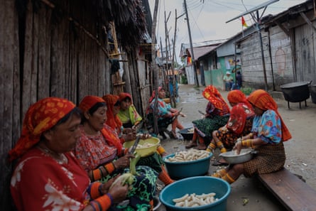 Women sit on low benches at the side of a road with peeling bananas into large bowls.