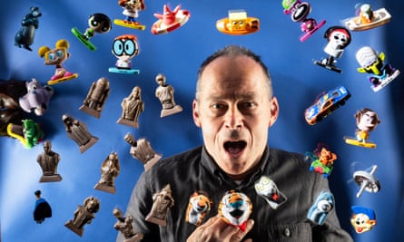 Ian Madeley holding a toy and with many other toys scattered on a blue background behind him