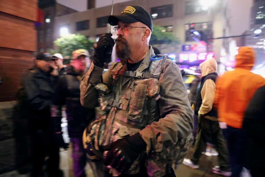 A man in military attire and a black baseball cap speaks into a radio as he surveys a crowd.