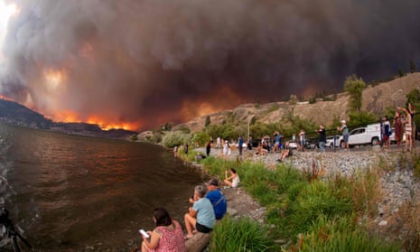 People sit by a creek with huge wildfire and billows of smoke in the background.