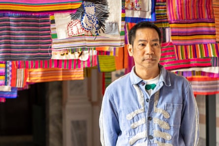 A south-east Asian man in an embroidered shirt stands in front of lots of small striped pieces of cloth hanging from the ceiling 