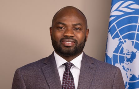 A young African man in a suit standing in front of a UN flag