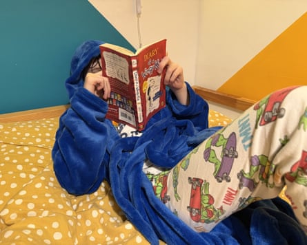 A child wrapped in a blue hooded blanket robe lies reading a book, testing gifts for 9 year olds