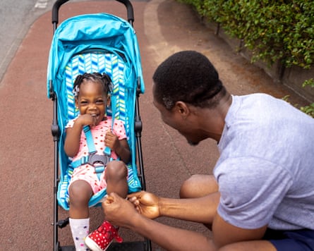 Father of toddler straps his daughter’s Velcro shoes as she sits in her pushchair while on a walk. She is giggling and smiling as her father interacts with her.