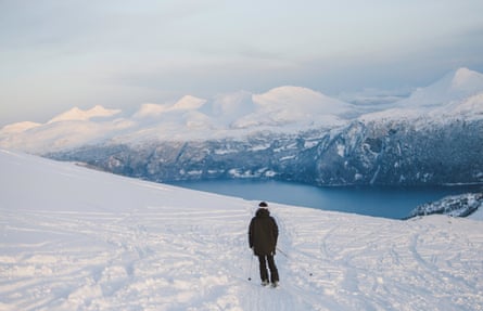 A lone skier on a snowy plateau looking at a fjord and mountains