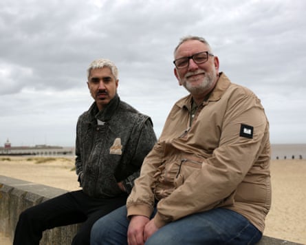 Azhar and Ivan sit together on a stone wall beside a sandy beach