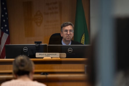 Brian McDonald, a judge with light-toned skin and wearing glasses, sits at a judge’s podium. A person wearing a peach-colored top and with light brown hair sits facing him.