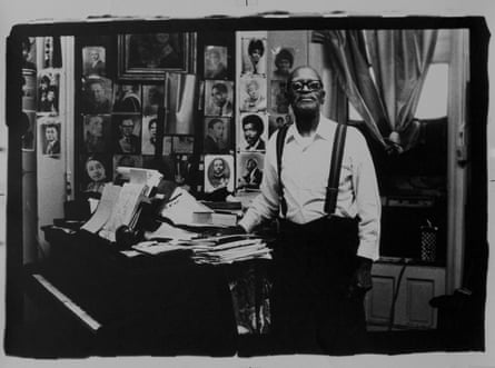 black and white photo of man next to piano stacked with papers, with framed portraits behind it