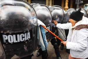 Police officers stand behind thick shields in a close line as an elderly woman in a white anorak and black woollen hat shouts at them; she is holding a pale blue and white Argentinian flag against the shield of one officer and looks angry
