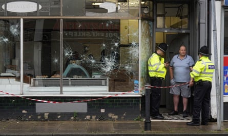 Two police officers talk to a man standing by smashed shop windows.