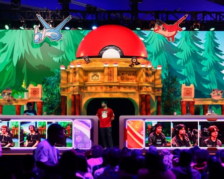 A person standing on a stage, with screens showing other people in front of a crowd, with Pokémon characters displayed in the background