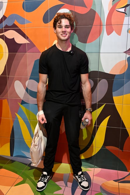 A man in black T-shirt and black jeans stands in front of a multicoloured background