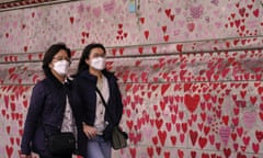Two women in face masks walk past a wall with many hearts drawn on it.