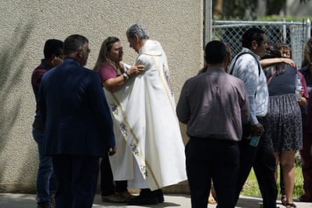 A priest in ceremonial robes hugs a woman as other mourners mill around.