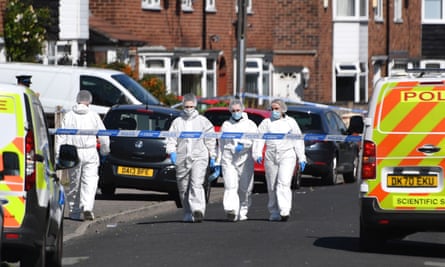 Three forensic officers in white overalls and masks walking down the centre of a residential street framed by two police vans, one on either side of the road