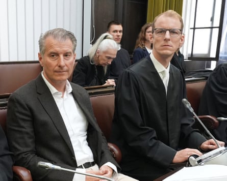 Stephan Hensel sits next to his lawyer in the court room; he has short grey hair and wears a white shirt and black jacket.