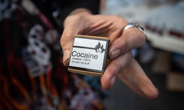 A close-up shot of a man's hands holding a small box labeled 'Cocaine - 100%, no cut / no buff.'