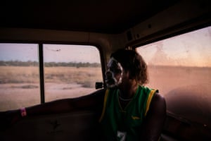 An Indigenous man with lines of white face paint and wearing a basketball jersey sits in the back of a car driving along a dirt road and sending up dust behind it