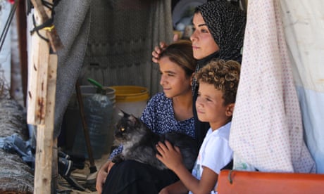 Bir kadın ve iki çocuk çadırlarından dışarı bakıyor, kadının kucağında bir kedi oturuyor