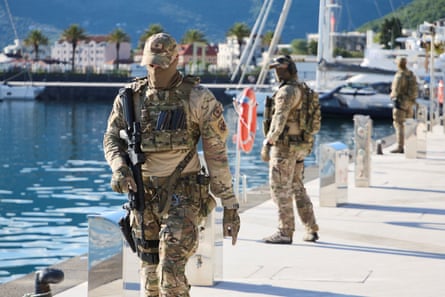 Armed personnel in camouflage, carrying guns, patrol on a harbourfront. There are yachts in the background.