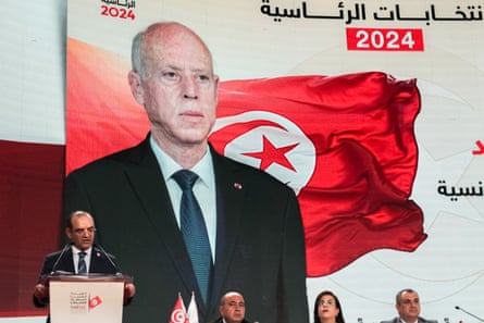 A man talks at a podium in front of a huge banner of an old Arab man standing in front of a red Tunisian flag
