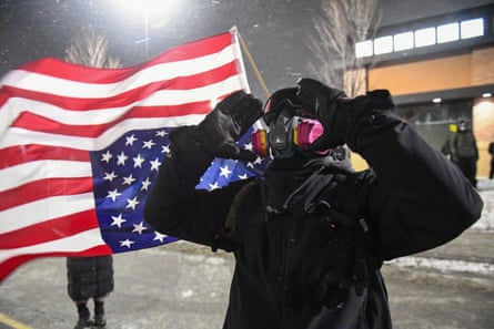 Person in all black, with face covered, in front of an upside down US flag