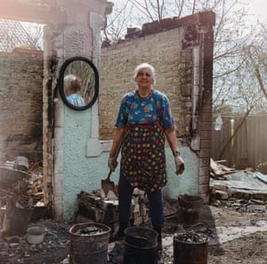 A white-haired woman stands in the ruins of a house, with three buckets and a small spade in her hand