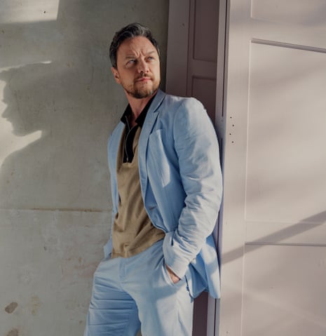 James McAvoy leaning against an open concertina door, wearing pale blue trousers and jacket, over a fawn shirt, with shadows