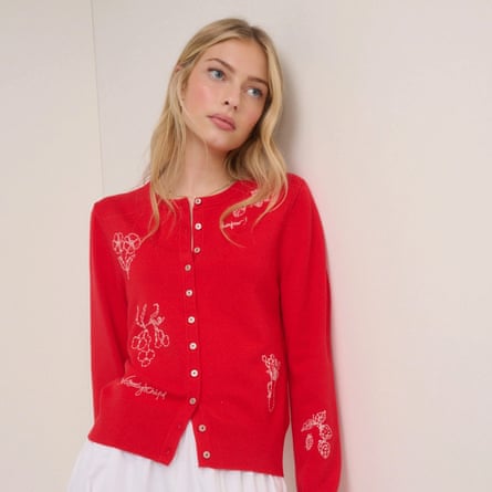 A model leans against a wall wearing a red cardigan with white floral and cherry embroidery