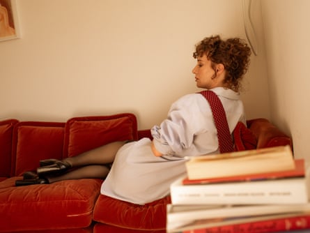 Portrait of Miranda July lying on a red sofa wearing an oversized shirt and tie that’s been left undone, with her back to the camera