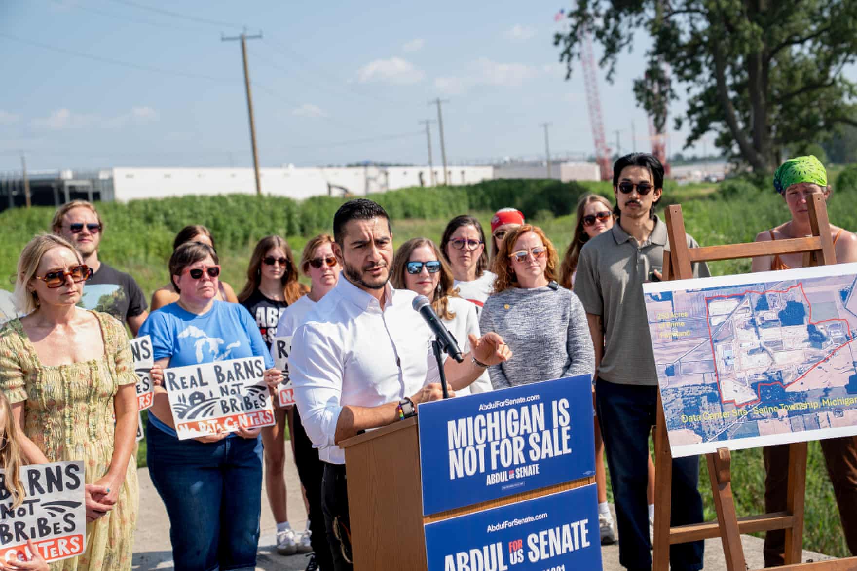 Man at a podium with people holding signs behind him and a boundary map on a stand
