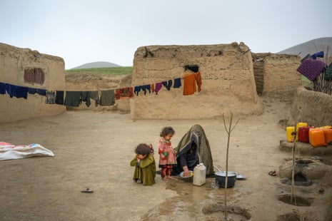 A woman crouches to wash dishes in a yard next to two small children. Washing is strung from the mud-brick house behind her.