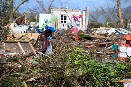 A woman is seen lifitng debris next to an almost entirely destroyed building, of which only one wall remains, with piles of possessions, trees and building materials