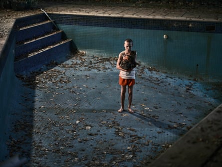 A young boy with a shaved head stands in an empty leaf-littered pool, holding a cat, looking sinister