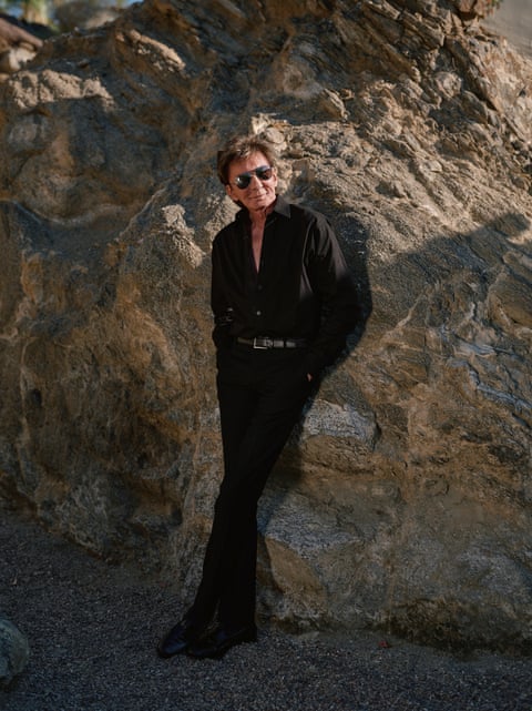 Barry Manilow in a black shirt, black trousers and sunglasses leans against some rocks.
